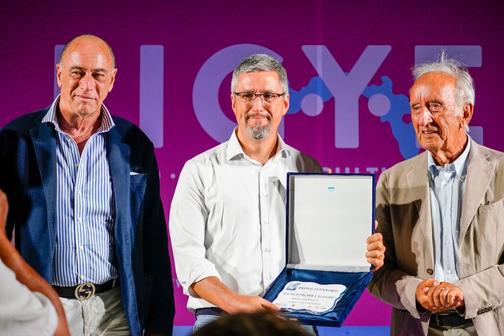 Paolo Malaguti mostra la targa del premio Alssio 100 Libri un autore per l'europa, accanto a lui il sindaco Marco Melgrati e Luigi Beccaria presidente della giuria del premio, sullo sfondo il logo del festival ligyes 2025